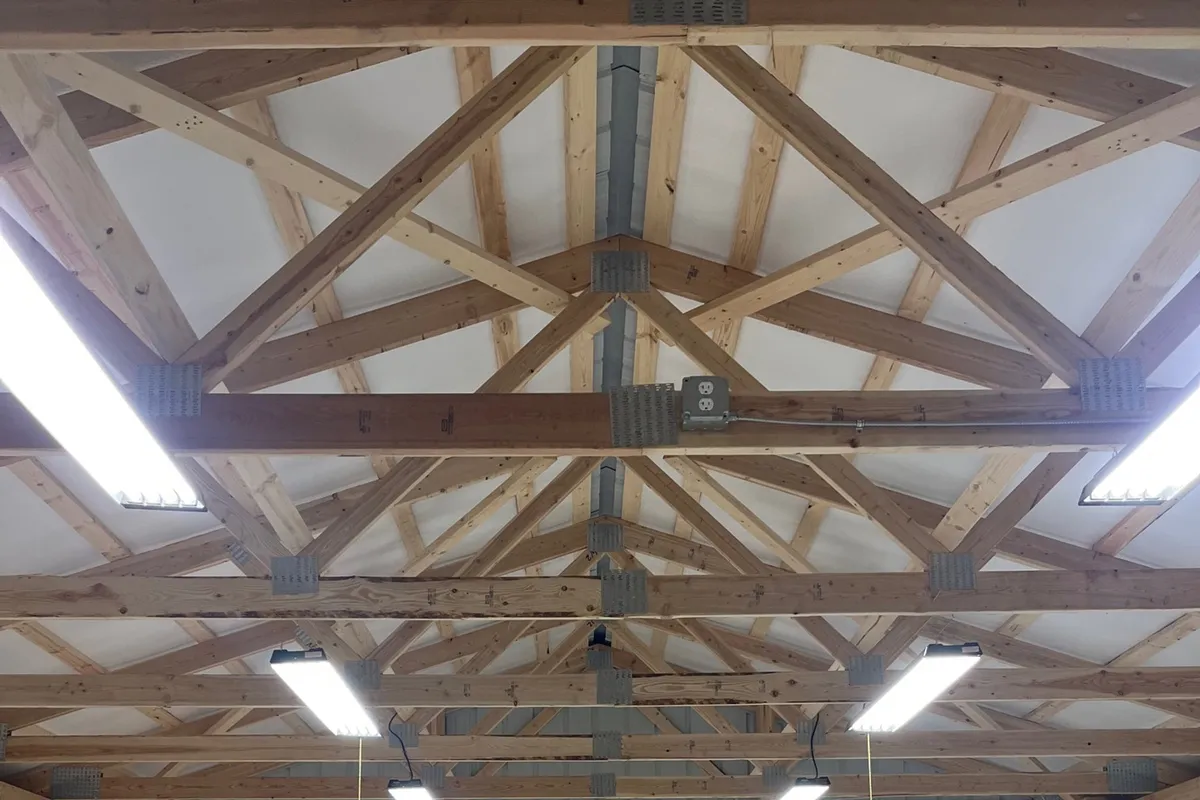 A wood-truss pole-building shop lit by a full grid of LED high-bay fixtures, with outlets on the posts and a workbench below.