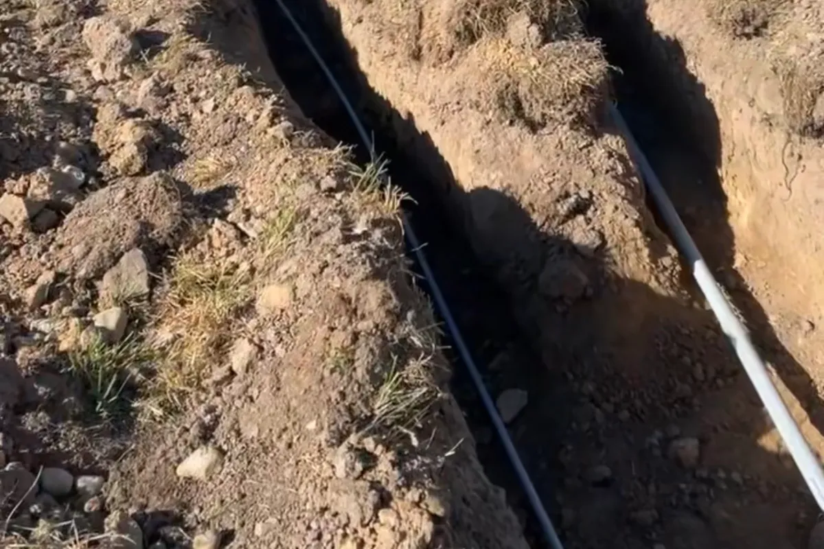 An open utility trench with buried conduit running across a rural field toward the Montana mountains.