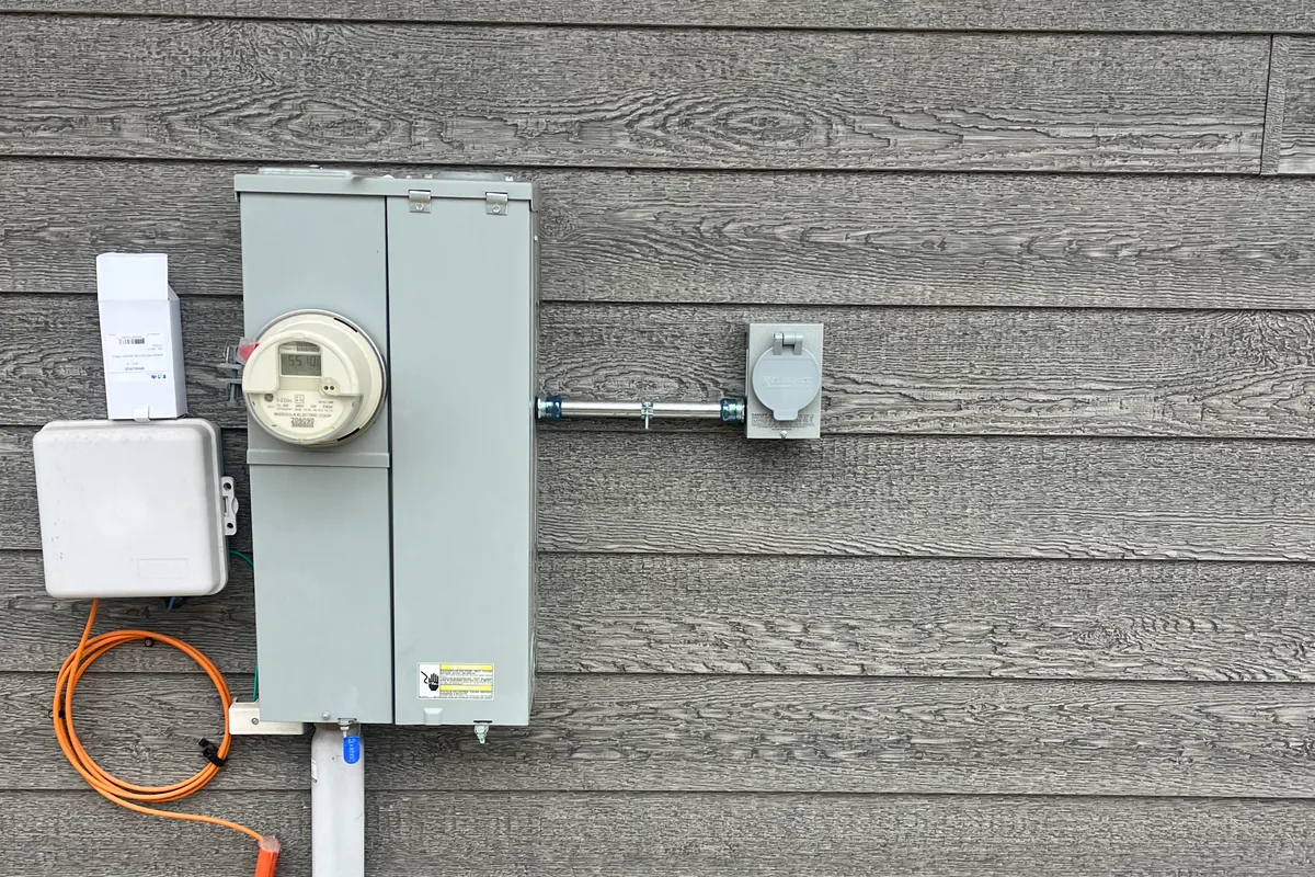An exterior electric meter and service riser conduit on a home, with a weatherproof outlet on gray siding.