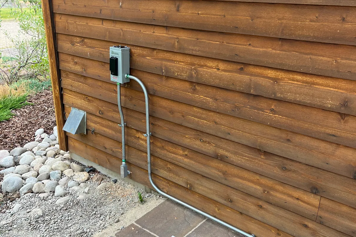 An exterior disconnect box fed by galvanized rigid conduit routed down the wood siding of a home to a weatherproof outlet.