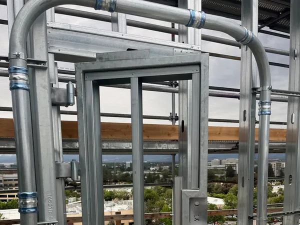 Large rigid conduit sweeps and compression couplings beside a rooftop catwalk gate on a commercial high-rise above the campus skyline.