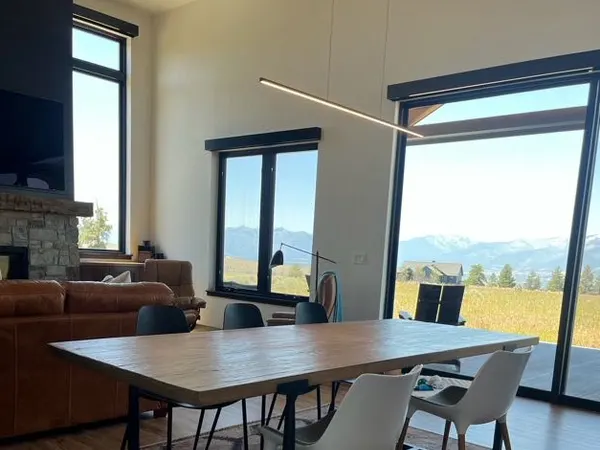 A modern great room lit by a slim linear LED pendant over the dining table, with floor-to-ceiling glass framing the Bitterroot mountains.