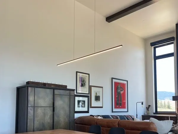 A linear LED pendant suspended over a dining table beneath a beamed ceiling, beside a gallery wall and valley-view windows.