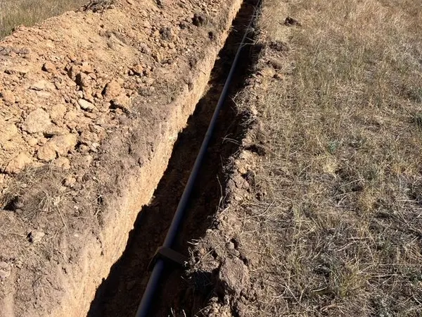 A long open trench with underground conduit running straight across a golden Bitterroot Valley field toward the tree line.