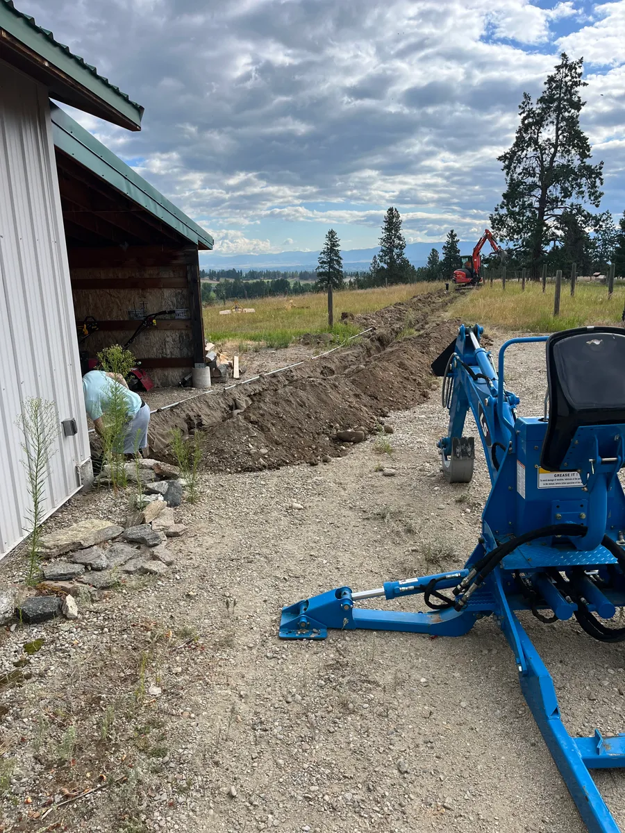 An open utility trench and a compact backhoe beside a metal building on a rural job site, with a worker and an excavator in the background.