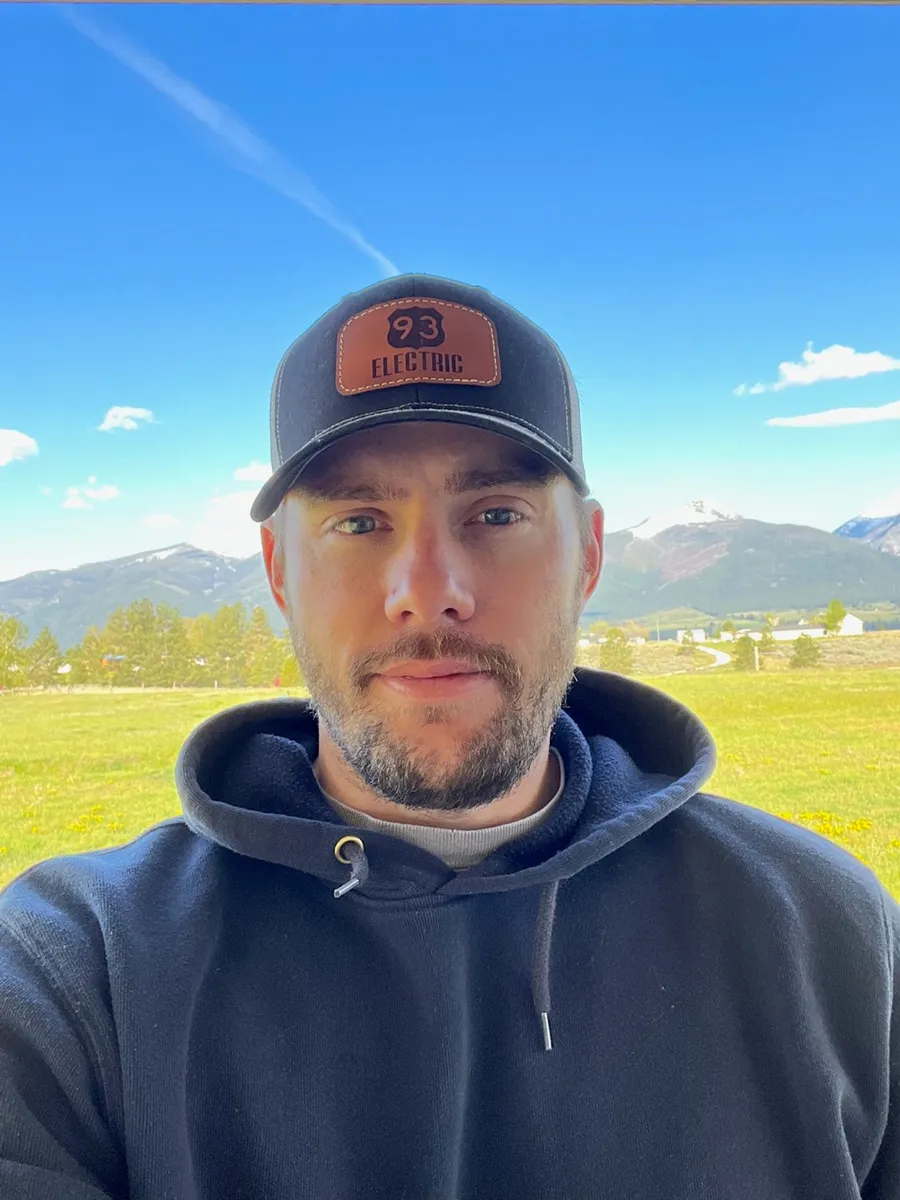 Josh Brown, owner and Master Electrician of 93 Electric, wearing a 93 Electric cap with the Bitterroot Valley mountains behind him.