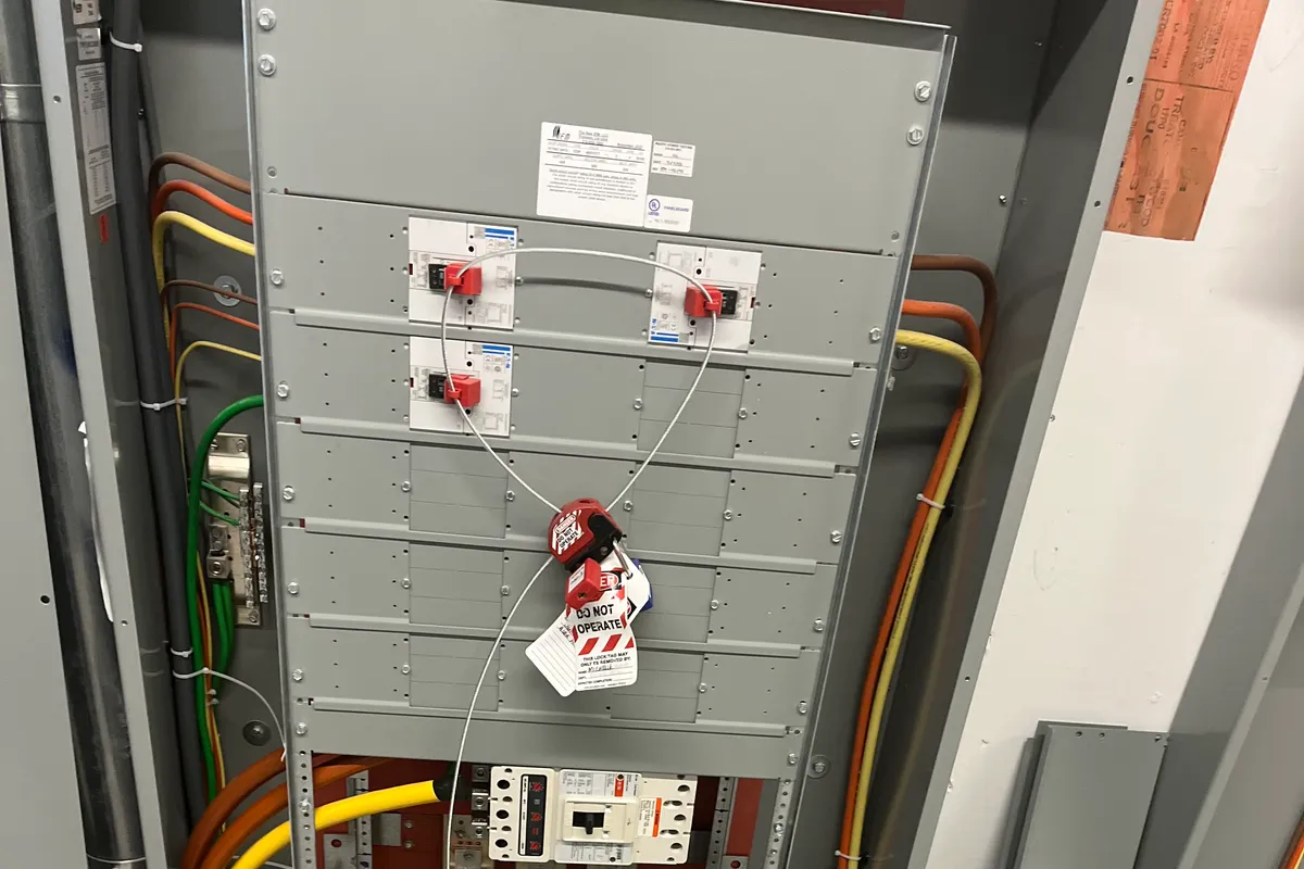 An open commercial power distribution panel with a red Do-Not-Operate lockout tag, color-coded feeder cables, and flexible conduit rising from a raised access floor.