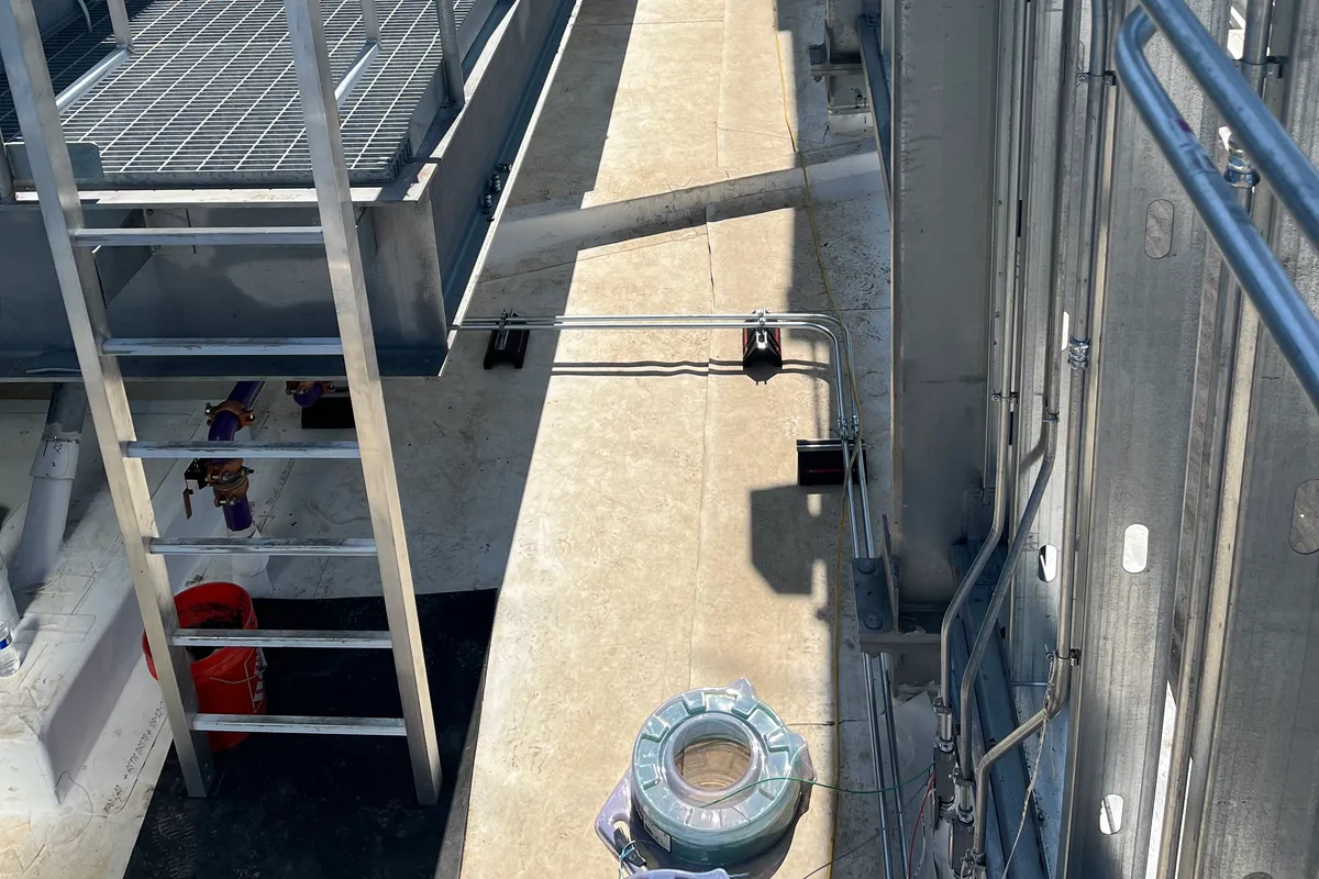 A galvanized EMT conduit run installed along a rooftop wall beside commercial HVAC units, with a steel-grating catwalk and access ladder.