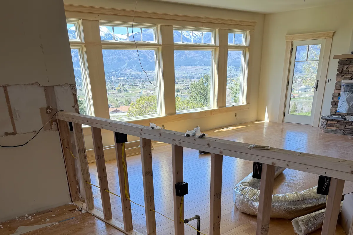 A home mid-remodel with a newly framed pony wall holding rough-in outlet boxes and cable, hardwood floor and mountain views through the windows.